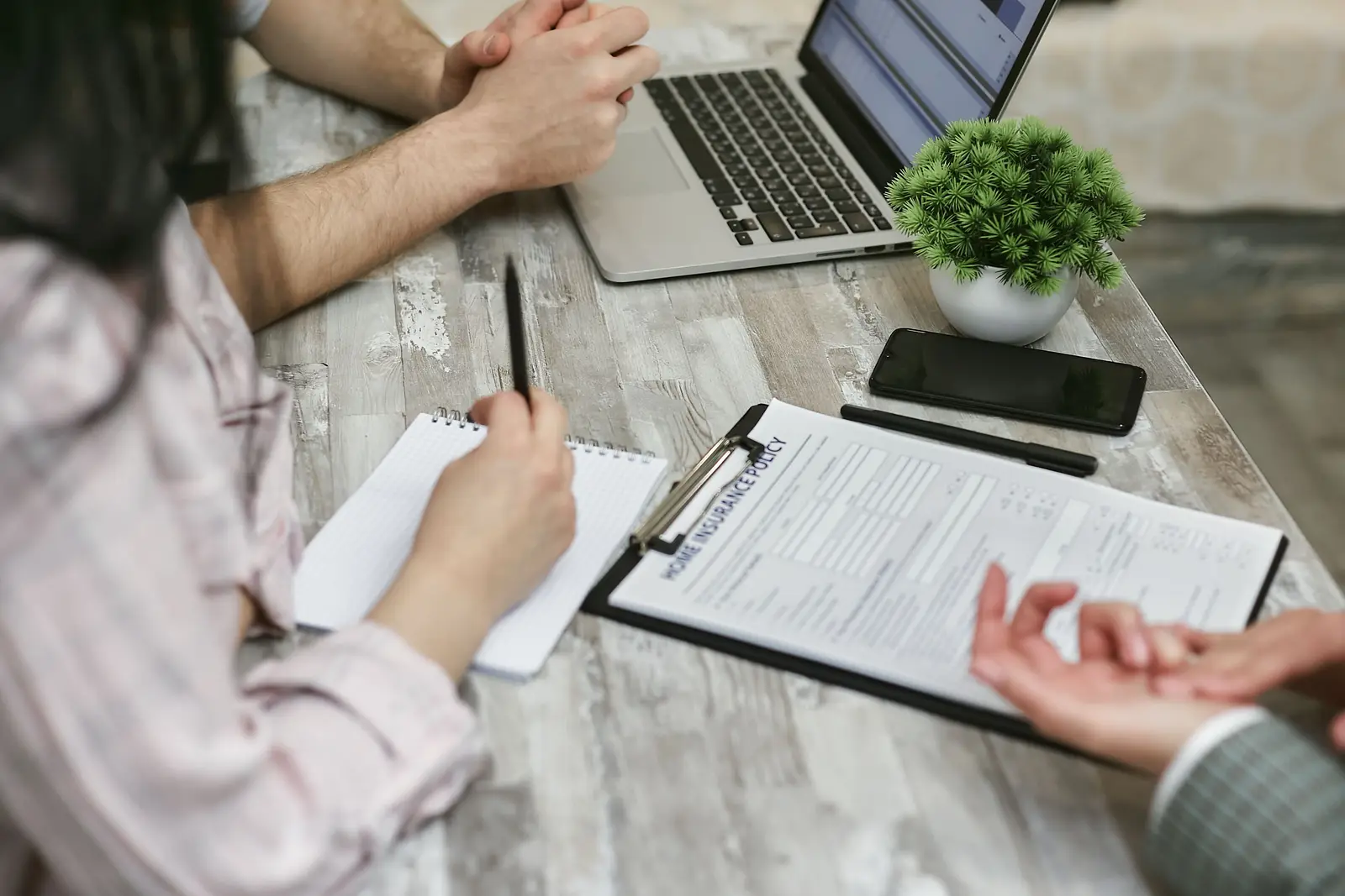 An agent and clients going over a home insurance policy with a pen, a laptop and a clipboard on the table